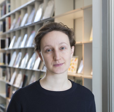 Femme aux cheveux courts debout devant des étagères remplies de livres dans une bibliothèque moderne, regardant l’objectif avec un léger sourire.
