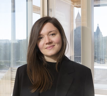 Portrait d’une femme souriante aux cheveux bruns, vêtue d’un blazer noir, debout près d’une baie vitrée offrant une vue sur la ville et une tour en arrière-plan