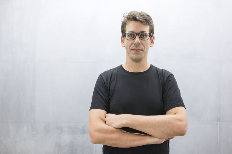 Homme aux cheveux courts et lunettes, debout devant un mur gris clair, bras croisés, regard assuré