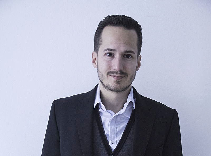 Homme barbu aux cheveux coiffés en arrière, portant un costume noir, un gilet gris foncé et une chemise blanche, posant devant un mur blanc uni.