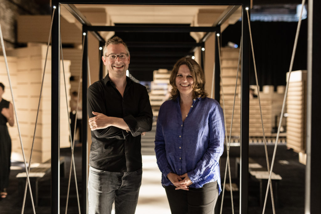 Deux personnes souriantes, un homme en chemise noire et lunettes et une femme en chemise bleue, posent devant un couloir d'installation en modules de bois clair suspendus par des câbles dans un espace d'exposition design.