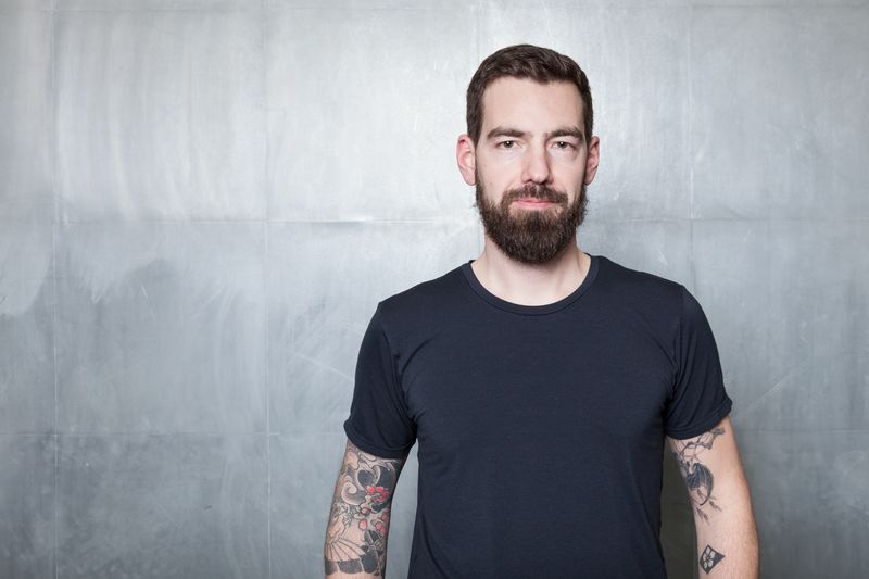 Portrait d’un homme barbu portant un t-shirt noir et arborant des tatouages colorés sur les avant-bras, debout devant un mur en béton gris