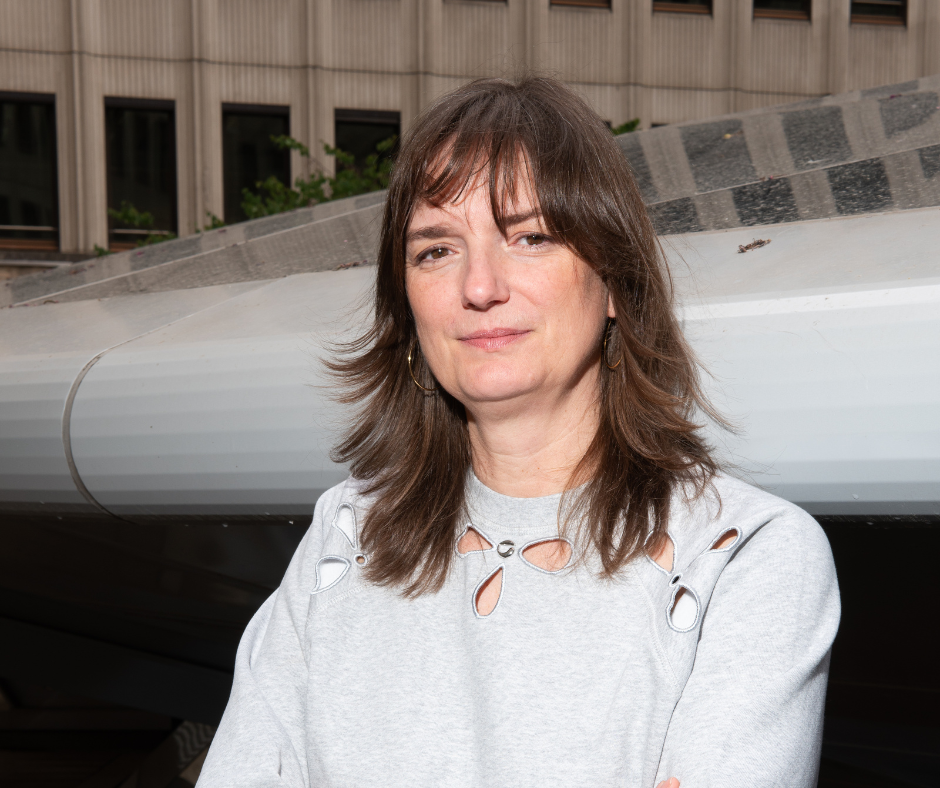 Portrait d’une femme brune vêtue d’un pull gris à découpes, bras croisés devant un bâtiment moderne en arrière-plan.
