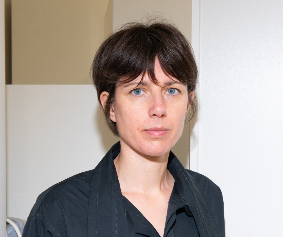 Portrait rapproché d’une femme au cheveux bruns courts et aux yeux bleus, visage sérieux, chemise noire et arrière-plan neutre blanc.