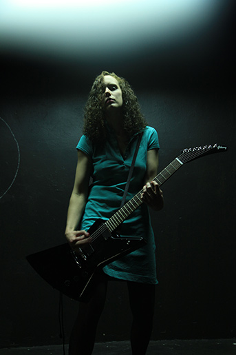 Femme guitariste en robe bleue jouant d’une guitare électrique noire sous une lumière tamisée sur une scène sombre